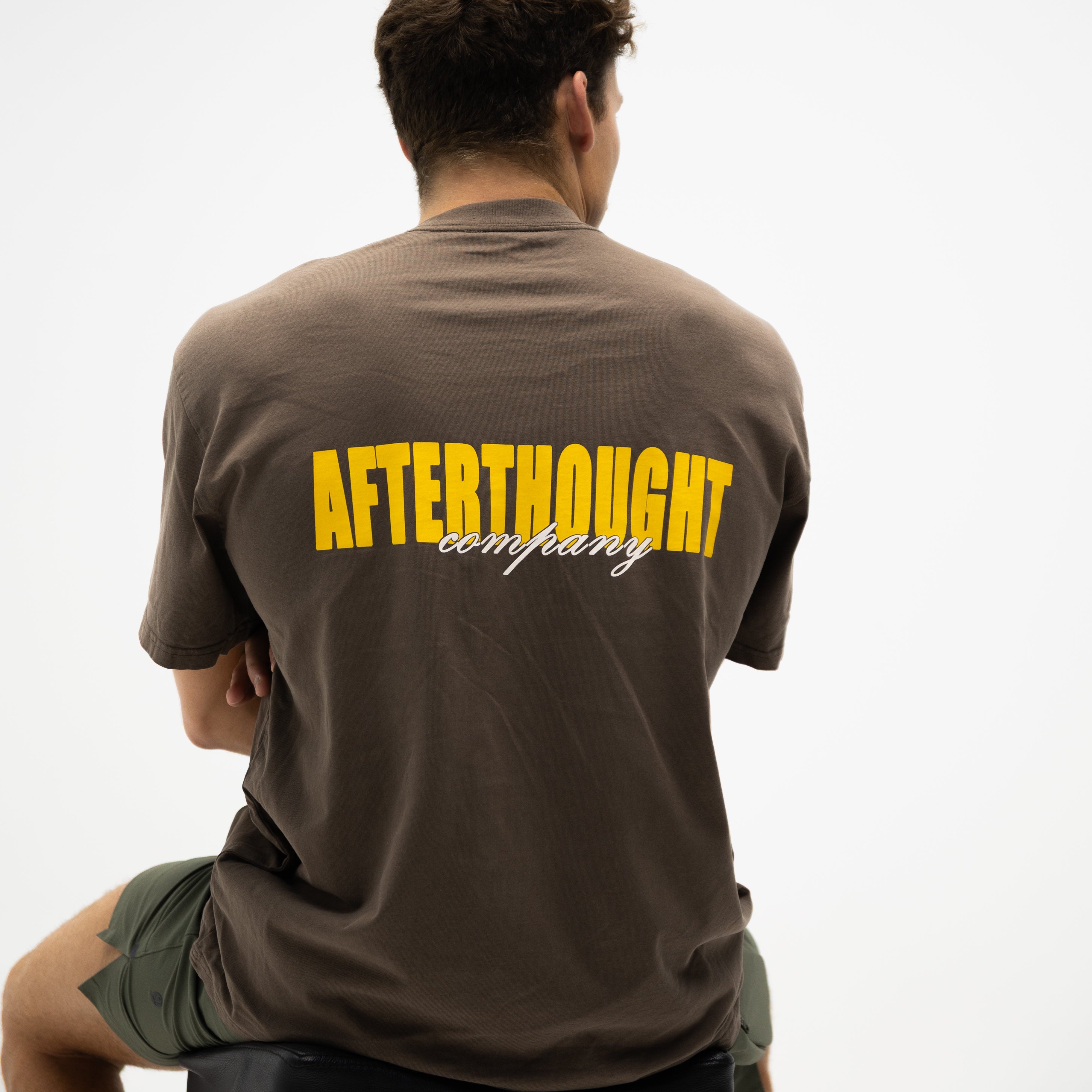 Person wearing a brown t-shirt with 'Afterthought Company' printed on the back against a white background